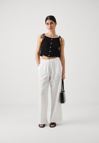 Crni pleteni crop top s prednjim gumbima i vezicama, uparen s bijelim lanenim hlačama širokih nogavica i crnim sandalama, držeći crnu torbu.