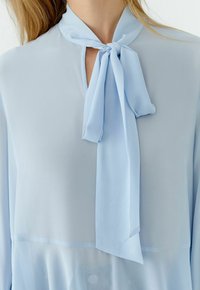 Hellblaue, transparente Bluse mit langen Ärmeln und einer Schleife am Ausschnitt, getragen von einer Person mit blondem Haar.