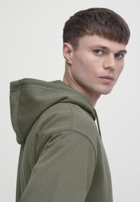 Oliivrohelise kapuutsiga dressipluus lõdva lõikega, pehme tekstuuriga ja rõhutatud õmblustega õlgadel ja kapuutsil.