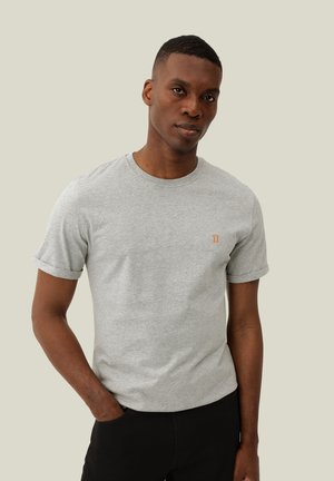 Homme portant un t-shirt gris clair à manches courtes avec un petit symbole orange sur la poitrine, debout avec une main dans la poche.