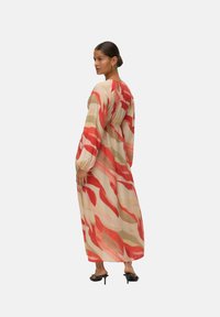 Femme portant une longue robe ample avec des rayures abstraites rouges, beiges et roses, se tenant de dos en partie tourné, chaussée de sandales à talons noires.