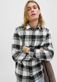 Młoda osoba z długimi blond włosami, ubrana w czarno-białą koszulę w kratę, z założonymi rękami, pokazująca tatuaże na przedramionach, stojąca na jednolitym tle.