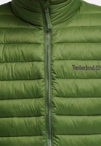 Grön puffjacka med hög krage, horisontell quiltad struktur och framdragkedja. Har Timberland-logotypen på vänster sida.