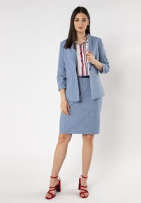 Lichtblauw pak van een textuurstof, bestaande uit een blazer en een kokerrok. Gepaard met een gestreept blouse en rode hoge hakken.