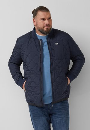 Mann mit Bart, der eine marineblaue gesteppte Jacke über einem blauen Sweatshirt und Jeans trägt, steht mit den Händen in den Taschen und schaut nach rechts.