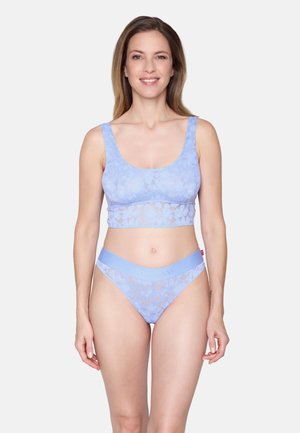 Femme souriante, portant un bralette en dentelle florale bleu clair et une culotte taille haute assortie, debout devant un fond blanc uni.