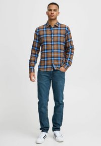 Camisa de cuadros marrón y azul de botones, hecha de tela, que presenta un cuello, mangas largas y un corte recto. Llevada con jeans oscuros y zapatillas.