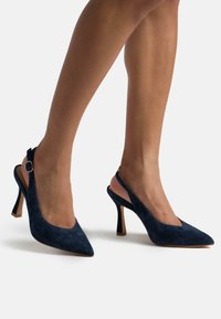Talons hauts slingback en velours bleu marine avec un bout pointu, un talon de 7 cm et un accent en boucle argentée sur la bride. Texture lisse avec un design minimal.