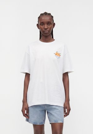 Femme aux cheveux tressés portant un tee-shirt blanc oversize avec un motif de fruit orange et un short en jean bleu clair, debout devant un fond uni.