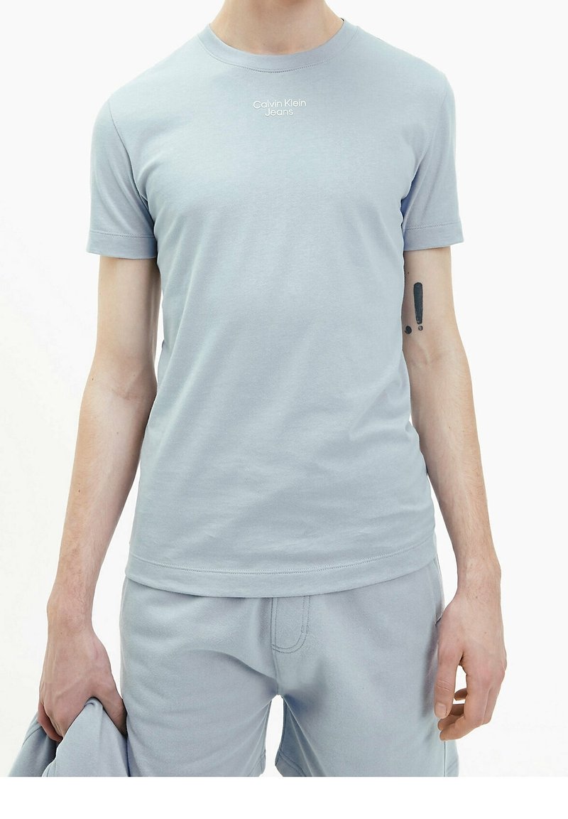 Mann trägt ein hellblaues Calvin Klein Jeans T-Shirt und passende Shorts, hält ein Kleidungsstück, mit Ausrufezeichen-Tätowierung am linken Arm.