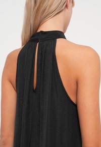 Top preto sem mangas com um pescoço alto, apresentando uma abertura em forma de fechadura nas costas e um tecido texturado. Design suave e leve.