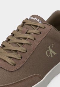 Κοντινό πλάνο καφέ αθλητικού παπουτσιού Calvin Klein με λεπτομέρειες από ύφασμα και σουέντ, μπεζ κορδόνια, λογότυπο "cK" στο πλάι και λευκή ελαστική σόλα.