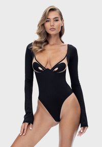 Kobieta z falującymi blond włosami, ubrana w czarny bodysuit z długim rękawem, który ma wycięcia i przezroczyste panele, stojąca na jednolitym jasnoszarym tle.