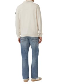 Jasnobeżowy, dziany sweter z prążkowanym kołnierzem, zestawiony z jasnoniebieskimi dżinsami. Sweter ma gładką fakturę i luźny krój.