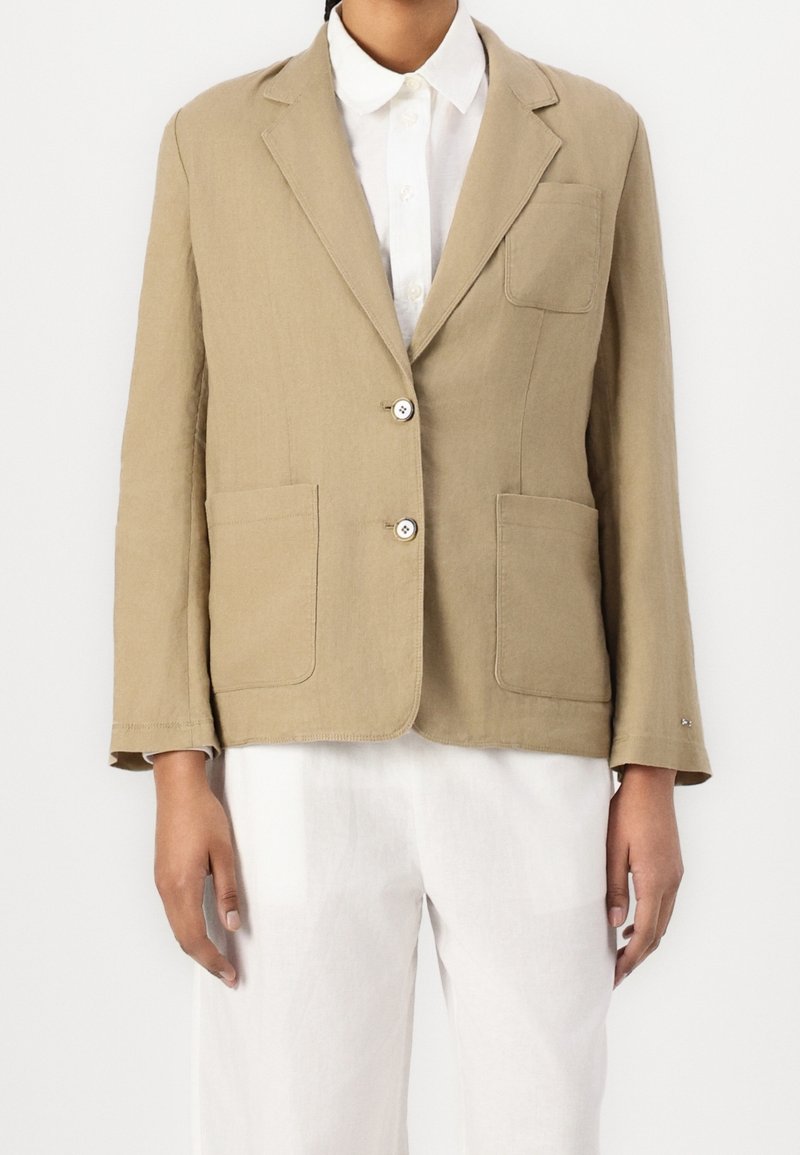 Blazer di lino beige con una tasca sul petto, due tasche frontali e chiusura con bottoni. Caratterizzato da un rever a scollo a V e maniche lunghe.