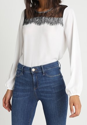 Vrouw draagt een witte blouse met lange mouwen met zwarte kanten details bovenaan en blauwe, hoog getailleerde spijkerbroek.