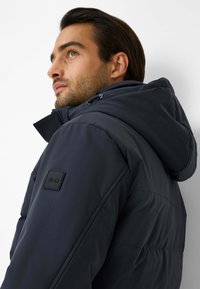 Giacca isolante blu navy con cappuccio, caratterizzata da una finitura testurizzata e una toppa nera con logo sul braccio; progettata per proteggere dal freddo.