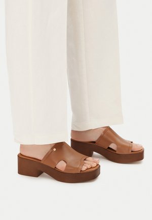 Sandales à plateforme en cuir marron à bout ouvert portées avec un pantalon blanc à jambes larges, mettant en valeur les pieds et les détails de la cheville sur un fond blanc.