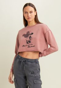 Sudadera corta rosa con gráfico negro de Minnie Mouse; textura suave, mangas anchas y dobladillo sin rematar. Combinada con pantalones cargo grises con logotipo.