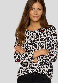 Woman with long brown hair wearing a patterned long-sleeve shirt with black, white, red, and purple shapes, arms crossed, standing against gray background.