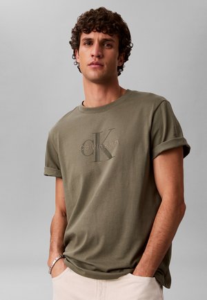 Uomo con capelli ricci che indossa una maglietta Calvin Klein verde oliva con maniche arrotolate e pantaloni beige chiaro, mani nelle tasche, sfondo neutro.