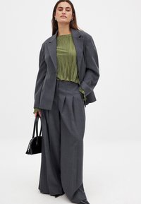 Grå skræddersyet blazer og vide bukser, kombineret med en plisseret grøn bluse. En sort håndtaske fremhæver outfittet. Glatte teksturer gennem hele looket.