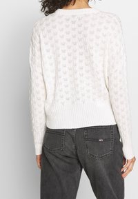 Weißer Strickpullover mit einem Rautenmuster, gerippten Bündchen und Saum. Kombiniert mit hoch geschnittenen, dunkelgrauen Jeans.