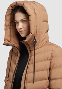 Jachetă puffer maro cu o glugă mare, având textură matlasată, șnur de strângere și fermoar frontal. Căptușeala de dedesubt este neagră și elegantă.