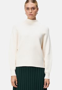 Pull en tricot côtelé couleur crème avec un col haut, des épaules tombantes et des manches longues, associé à une jupe plissée verte foncée.