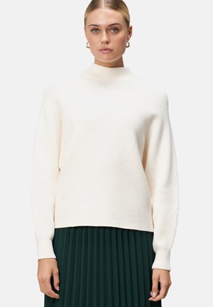 Pull en tricot côtelé couleur crème avec un col haut, des épaules tombantes et des manches longues, associé à une jupe plissée verte foncée.