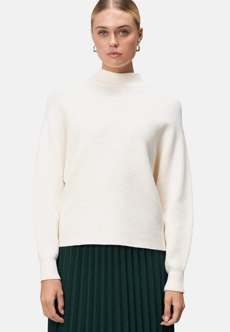 Pull en tricot côtelé couleur crème avec un col haut, des épaules tombantes et des manches longues, associé à une jupe plissée verte foncée.