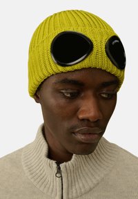 Berretto lavorato a maglia giallo con grandi occhiali neri sul davanti. Tessuto a coste con una vestibilità aderente. Indossato con un maglione grigio chiaro.