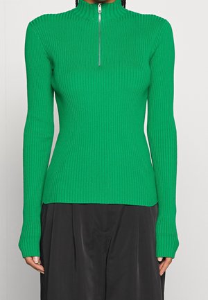 Femme portant un pull vert côtelé ajusté à manches longues avec une fermeture éclair argentée sur le devant et un pantalon noir à jambes larges.