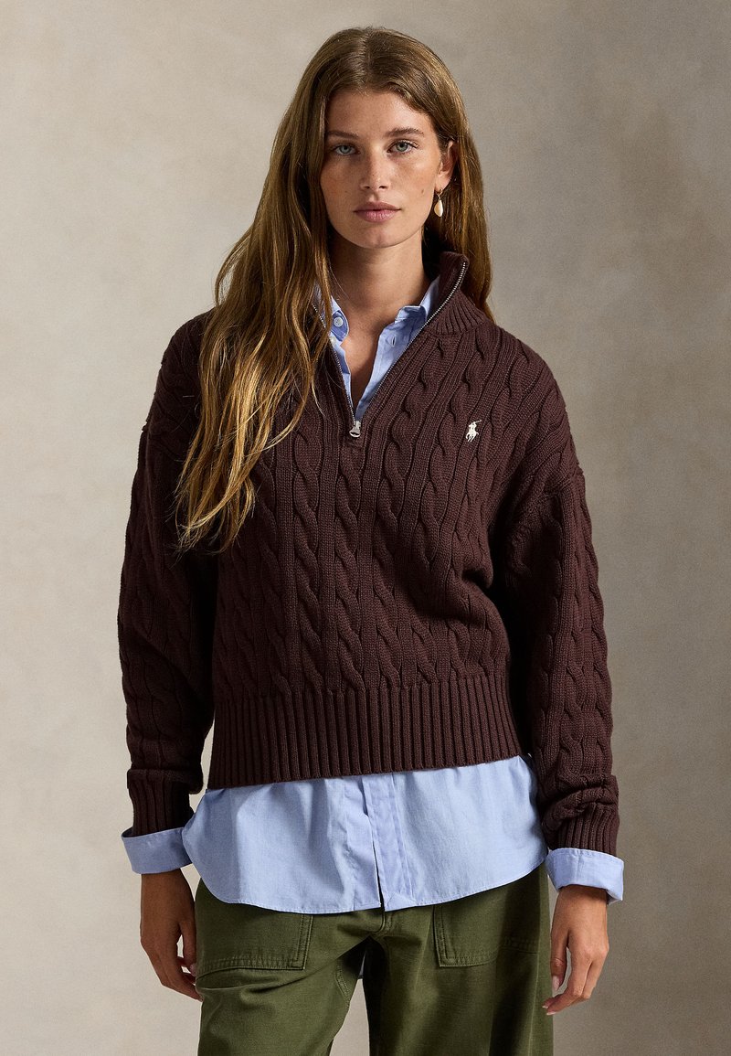 Hnedý sveter s vrkočovým vzorom, vysokým golierom, zapínaním na zips, rebrovaným lemom, nosený cez svetlomodrú košeľu s golierom a zelené cargo nohavice.