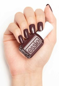 Vernis à ongles bordeaux foncé dans un flacon en verre avec un capuchon blanc, tenu dans une main avec des ongles manucurés de la même couleur.