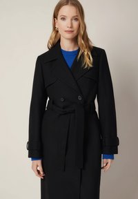 Czarny dwurzędowy płaszcz o szerokich klapach, z paskiem w talii i mankietami z regulacją. Noszony na niebieskim swetrze, wyeksponowany na neutralnym tle.