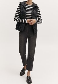 Musta tikattu liivi mustavalkoisen raidallisen pitkähihaisen paidan päällä, yhdistetty korkeavyötäröisiin tummiin farkkuihin ja mustiin loafereihin.