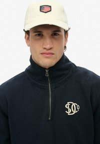 Marinefleece pullover med høj krave og halvlås, med et cremefarvet logo på brystet. Matchet med en cremefarvet kasket og broderet mærke.