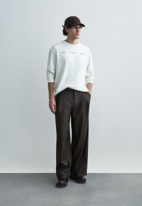 Camisola branca de mangas compridas com design de texto, combinada com calças largas castanho-escuras e ténis pretos. O conjunto é complementado por um boné castanho.