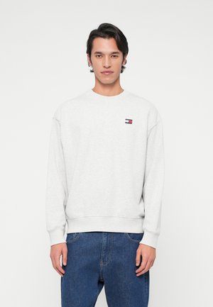 Man staat in een lichtgrijze sweatshirt met een klein rood, wit en zwart logo op de borst en blauwe jeans tegen een witte achtergrond.