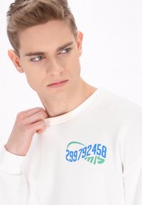 Sweatshirt in cotone bianco con scollatura rotonda. Design stampato in blu e verde con i numeri "299792458" e "mls". Texture morbida.