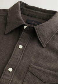 Barna gyapjú gombos ing, hegyes gallérral �és elülső zsebbel, három fehér gombot és egy fekete belső címkét mutat.