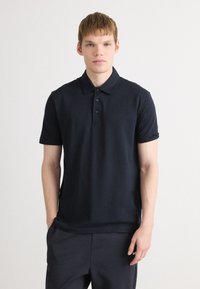 Joven con piel clara y cabello corto, vistiendo una camiseta polo oscura de color navy y pantalones a juego, contra un fondo blanco.