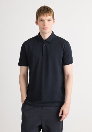 Young man with light skin and short hair wearing a plain dark navy polo shirt and matching pants against a white background.