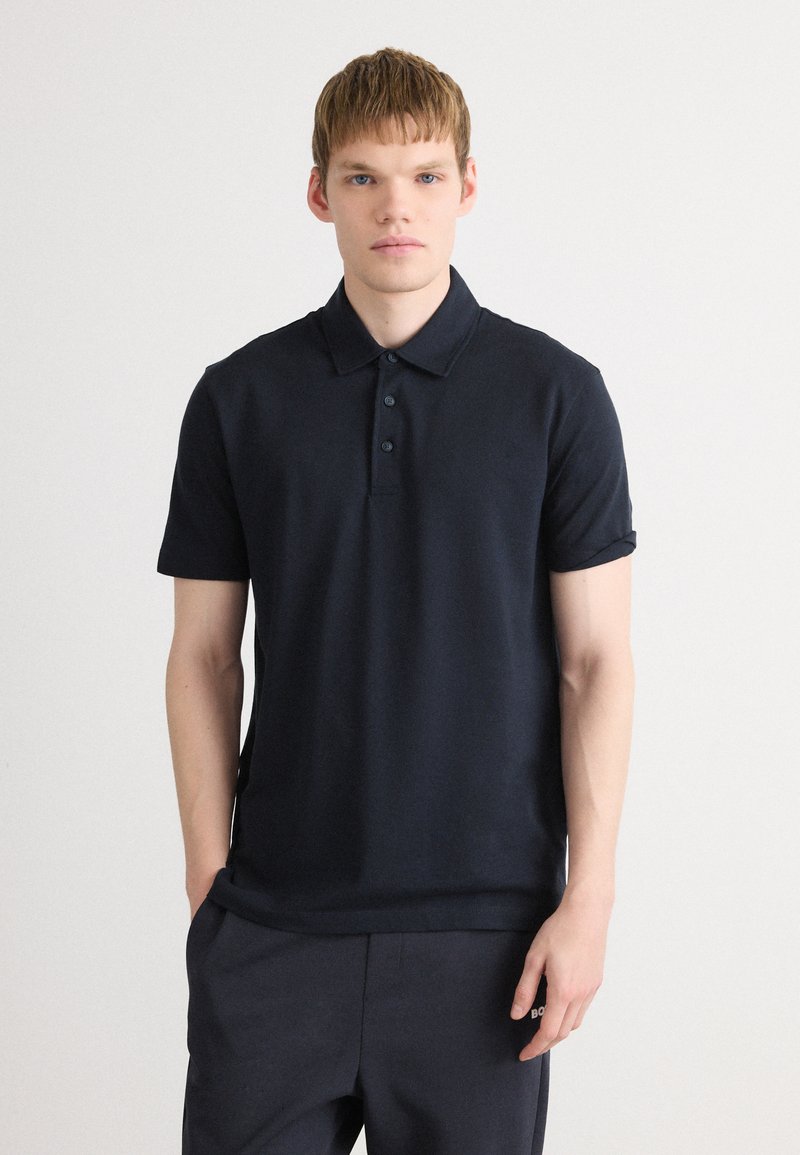 Joven con piel clara y cabello corto, vistiendo una camiseta polo oscura de color navy y pantalones a juego, contra un fondo blanco.
