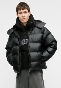 Črn pufi plašč s sijočim zaključkom, prevelike oblike, zadrgo in elastičnimi manšetami. Ima kapuco in subtilen detajl logotipa.