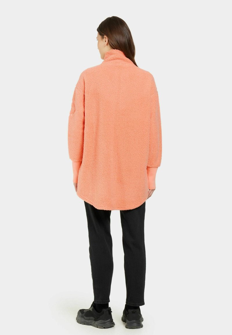 Flauschiger, korallenfarbener Pullover mit hohem Kragen und überschnittenen Schultern, kombiniert mit schwarzen, eng anliegenden Hosen und schwarzen Sneakern. Strukturiertes Material.