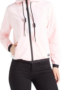 Jasnoróżowa kurtka przeciwdeszczowa z kapturem, przednim zamkiem błyskawicznym, czarnymi sznurkami do ściągania i elastycznymi mankietami, w zestawie z czarnymi dopasowanymi spodniami.