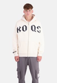 Cream hooded sweatshirt with black geometric print "KO QS" on front, zip closure, and front pockets. Ribbed cuffs; paired with gray cargo pants.