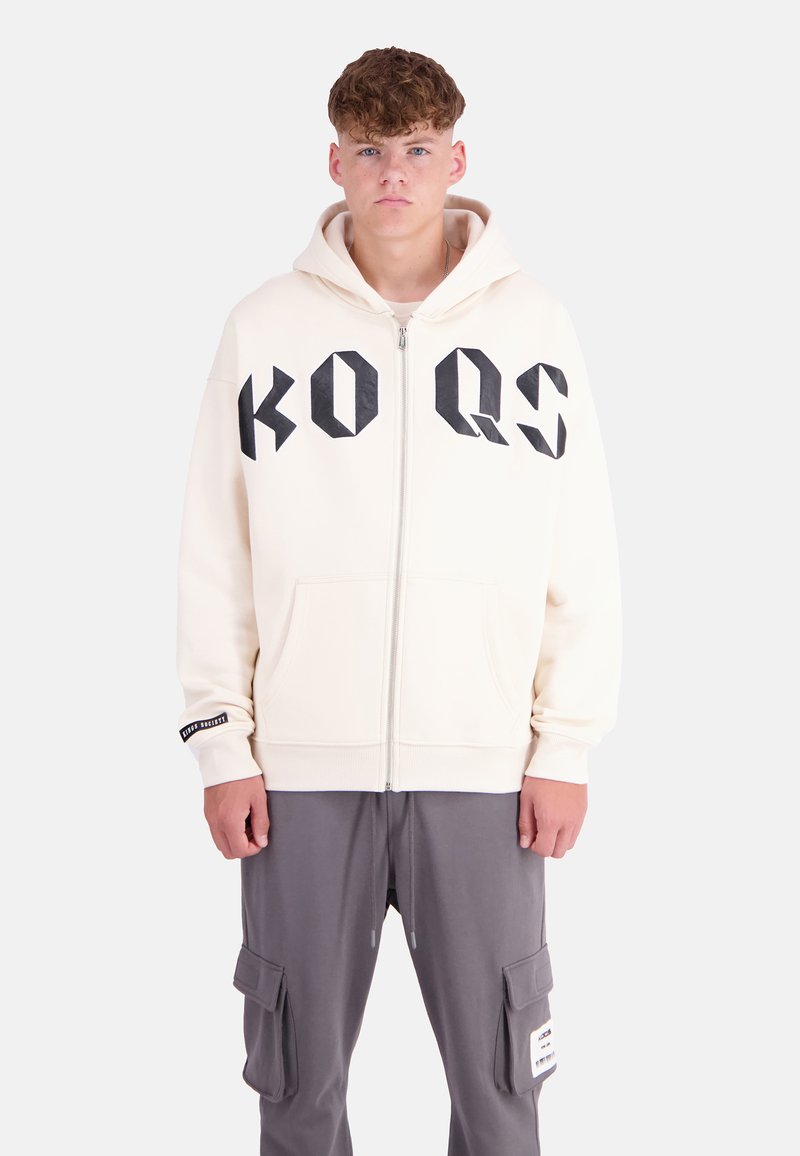 Cream hooded sweatshirt with black geometric print "KO QS" on front, zip closure, and front pockets. Ribbed cuffs; paired with gray cargo pants.